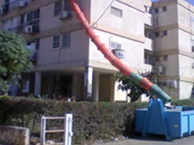 שרוולי פסולת כתום וירוק יורד מקומה גבוהה בבניין מגורים לתוך מכולה כחולה, לצורך פינוי פסולת בנייה